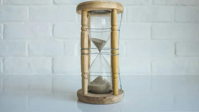 Reloj de arena de madera con la arena cayendo, sobre una mesa blanca frente a una pared de ladrillo