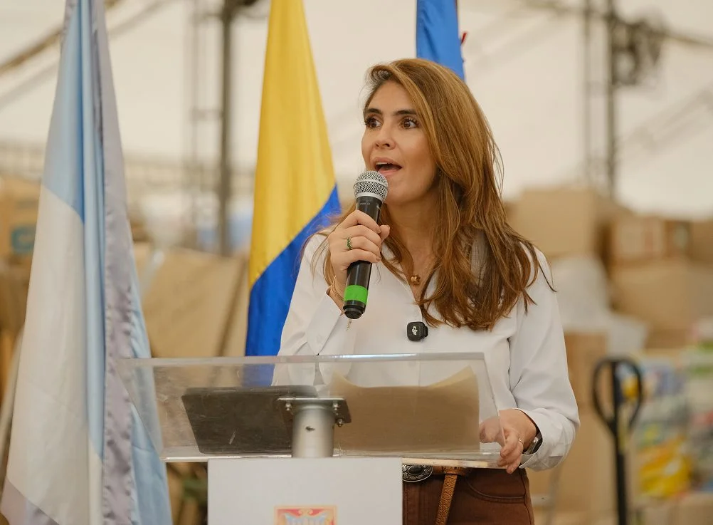 Una vocera interviene desde el atril durante el lanzamiento de la alianza, con cajas de insumos al fondo