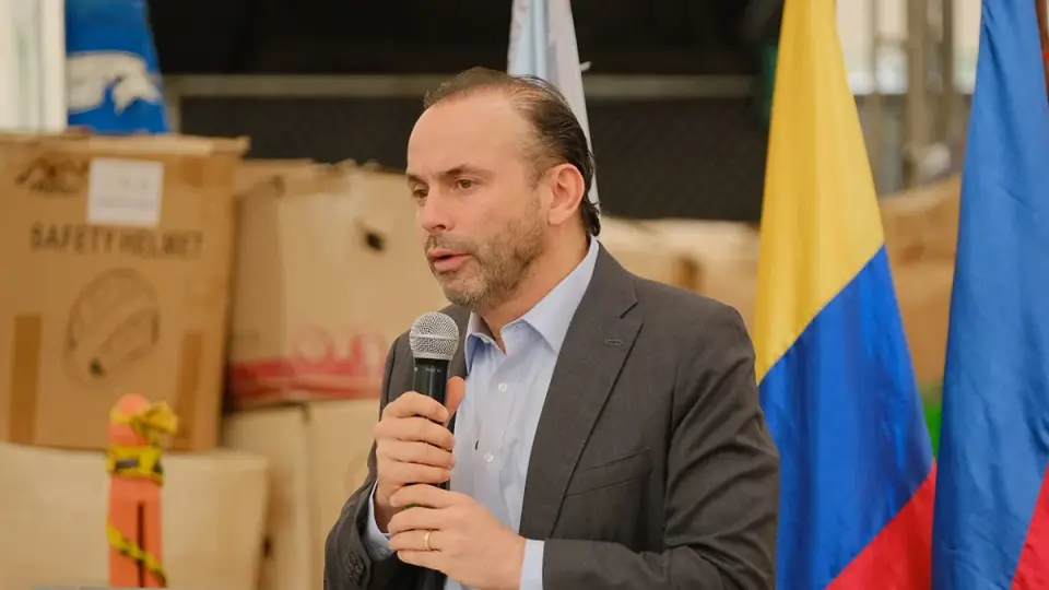 Un vocero habla con micrófono en un atril, frente a cajas de insumos y cascos de seguridad y junto a banderas de Colombia y Cali