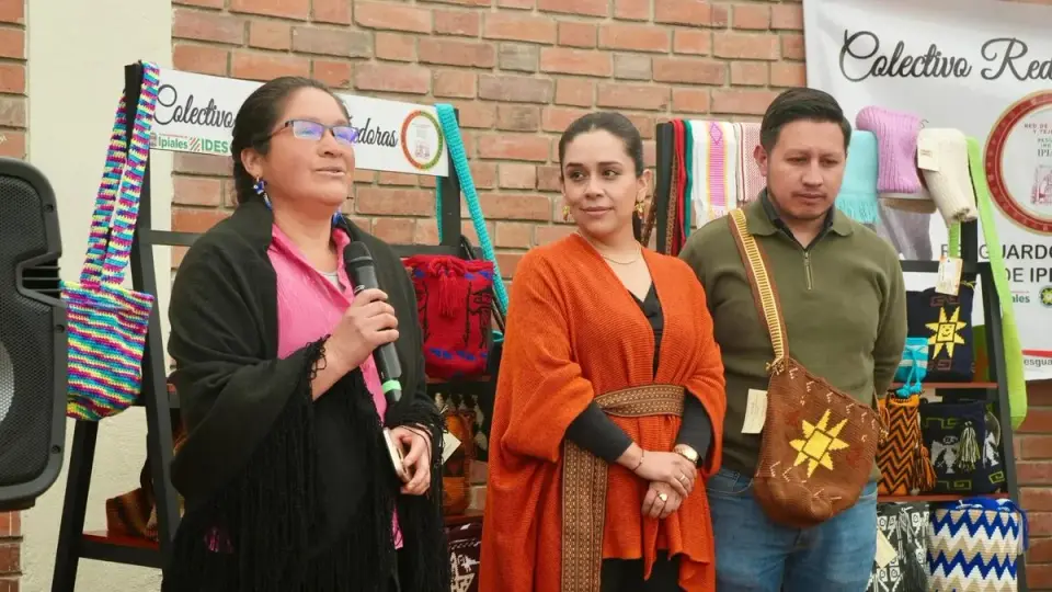 Una artesana del colectivo Red de Tejedoras habla con micrófono frente a estantes de mochilas y bufandas tejidas durante el lanzamiento de la ruta
