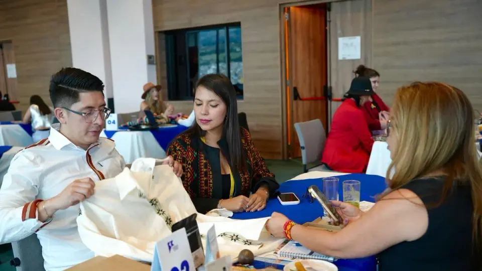 En una mesa de negocios, un empresario despliega una camisa bordada ante una compradora mientras otra participante observa